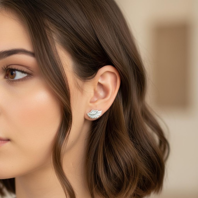 May include: A pair of silver-colored wing-shaped earrings. The earrings are worn on the ear of a person with brown hair. The earrings have a detailed feather design and are a stylish accessory.