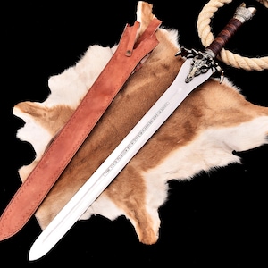 May include: A decorative sword with a silver blade and a brown leather-wrapped handle. The hilt has an animal head design. The sword lies on a brown and white fur-like surface, with a brown leather sheath. The sword has engraved text on the blade.