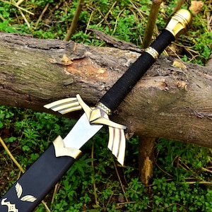 May include: A decorative sword with a silver blade and gold-coloured accents. The hilt is wrapped in black material, and the scabbard is black with gold-coloured leaf designs. The sword rests on a log in a natural outdoor setting.