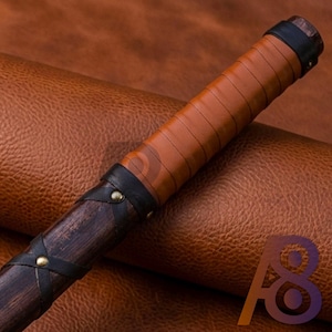 Könnte beinhalten: Nahaufnahme eines Holzstabs mit lederumwickeltem Griff. Der Griff ist mit braunem Leder umwickelt und hat schwarze Lederakzente und Messingnieten. Der Stab hat einen dunkelbraunen Holzschaft.