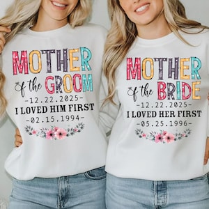 Op de afbeelding: Twee witte sweatshirts met de tekst "MOTHER of the GROOM" en "MOTHER of the BRIDE" in kleurrijke letters. Elk shirt heeft een datum en een bloemenontwerp. De sweatshirts worden gedragen met een blauwe jeans.