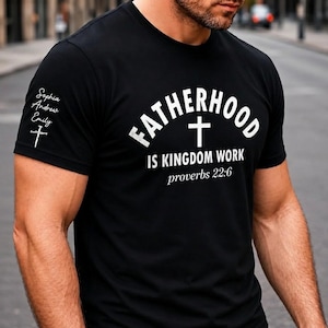 Op de afbeelding: Een zwart T-shirt met korte mouwen en witte tekst met de tekst FATHERHOOD IS KINGDOM WORK proverbs 22:6 en een kruisicoon. De rechtermouw toont de namen Sophia, Andrew, Emily en een kruissymbool gedrukt in witte inkt.