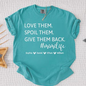 May include: A turquoise t-shirt with white text that reads "LOVE THEM. SPOIL THEM. GIVE THEM BACK. #mimilife" followed by names. The shirt has a crew neck and short sleeves. A gold necklace is visible.