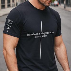 May include: Black t-shirt with white text that reads "fatherhood is kingdom work" and "PROVERBS 22:6". The sleeve has the names "Sophia, Andrew, Emily" and a cross. The shirt is a crew neck.