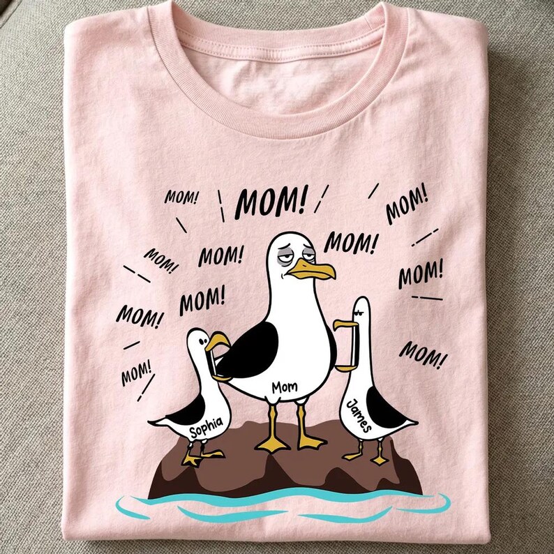 May include: Pale pink t-shirt with a cartoon seagull family illustration. The central seagull, labelled "Mom", is surrounded by speech bubbles saying "MOM!". The other seagulls are named "Sophia" and "James".
