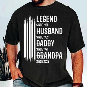 May include: Black t-shirt with white text that reads "LEGEND SINCE 1961, HUSBAND SINCE 1989, DADDY SINCE 1991, GRANDPA SINCE 2025". The shirt has a vertical white stripe design on the left side.