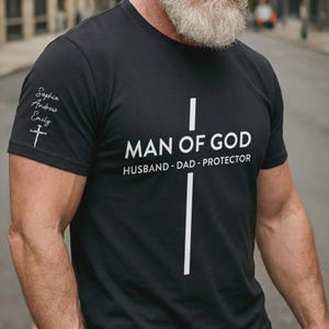 May include: A black short-sleeve t-shirt featuring a large white cross graphic on the chest with the text MAN OF GOD HUSBAND - DAD - PROTECTOR. The sleeve displays the names Sophia, Andrew, and Emily in white script above a small cross icon.