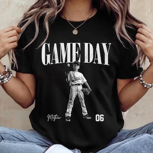 May include: Black t-shirt with the words "GAME DAY" in large white letters. A black and white image of a baseball player is centered below the text. The player is wearing a baseball uniform and holding a baseball and glove. The name "Matthew" and the number "06" are printed below the image.