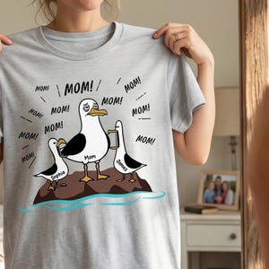 May include: A light grey t-shirt with a cartoon design of three seagulls on a rock, with the words "MOM!" and "MOM! MOM!" around them. The seagulls are holding phones, and the middle one has a sad expression. The names "Sophia", "Mom", and "James" are also visible.