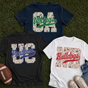 Pode incluir: Três t-shirts de manga curta em azul-marinho, preto e branco. Cada uma apresenta letras grandes com um padrão de camuflagem e o nome de uma equipa em escrita cursiva: Vikings em verde, Panthers em azul e Bulldogs em vermelho. Uma bola é visível.