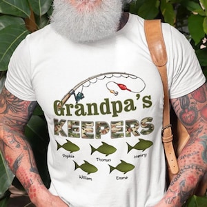 May include: White t-shirt with a fishing rod graphic and the text "Grandpa's Keepers" in green and camouflage lettering. Below the text are six green fish with names: Sophia, Thomas, Henry, William, and Emma.