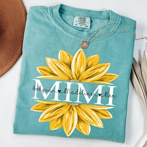 Peut inclure: Un t-shirt turquoise Comfort Colors avec un grand motif de tournesol jaune. Le mot "MIMI" est centré, avec les noms "Sophia, Madeline et Ben" en dessous. Un collier en or avec un pendentif est drapé sur le t-shirt.