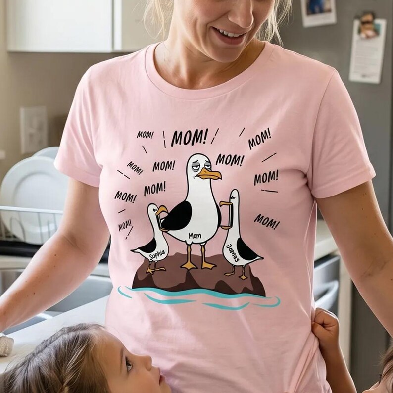 May include: A pale pink t-shirt with a cartoon seagull family design. The central seagull, labelled "Mom," looks stressed, with "MOM!" exclamations around it. Two smaller seagulls, labelled "Sophia" and "James," are also present. The design is a humorous depiction of motherhood.
