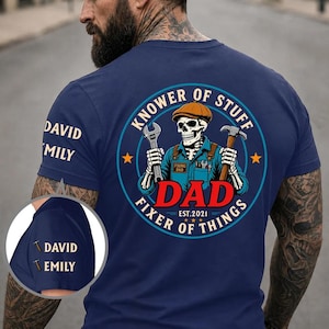 May include: A navy blue t-shirt featuring a circular graphic on the back with a skeleton wearing a cap, holding a wrench and hammer. The text reads "KNOWER OF STUFF", "FIXER OF THINGS", "DAD", "EST. 2021". The sleeve displays the names "DAVID" and "EMILY".