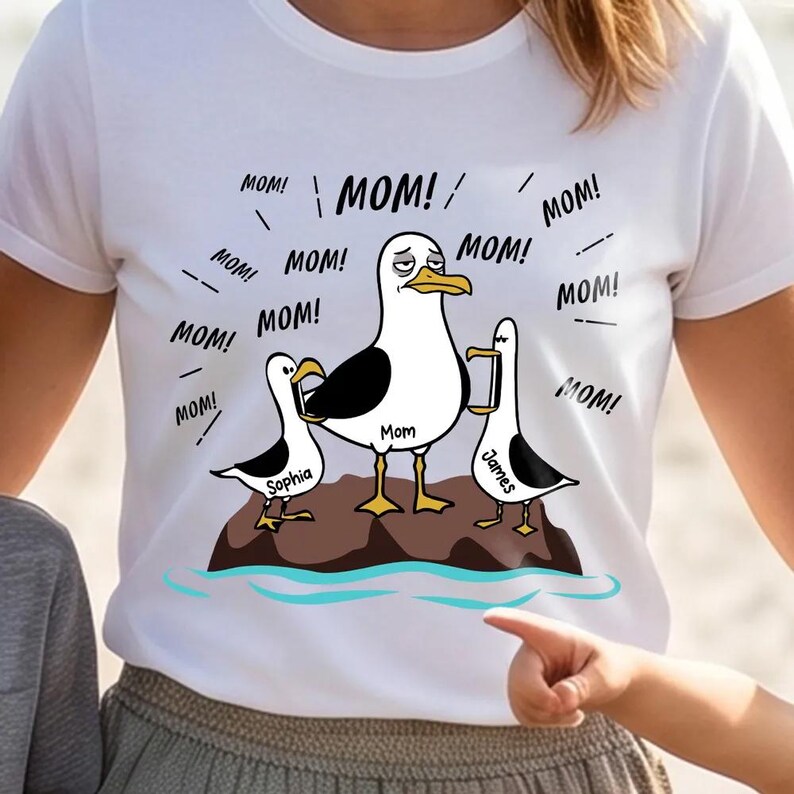 May include: White t-shirt with a cartoon seagull family on a rock, with the word "Mom" on the central seagull. The shirt has the text "MOM!" repeated around the seagulls, with the names "Sophia" and "James" on the smaller seagulls.