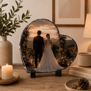 May include: A heart-shaped slate plaque featuring a printed photograph of a couple standing at an outdoor wedding altar during sunset. The plaque has a rough, chiseled edge and is supported by two small black stands on a wooden surface.
