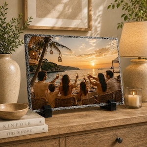 May include: A rectangular slate photo plaque featuring a printed image of a group of people sitting at a table on a beach at sunset, raising glasses in a toast. The plaque has a rough, textured edge and is supported by two black stands.
