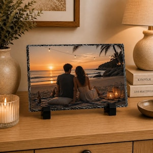 May include: A rectangular slate photo plaque featuring a printed image of two individuals sitting on a sandy beach at sunset. The scene includes a calm ocean, palm trees, string lights, and two glowing lanterns. The plaque has a rough, chiseled edge.