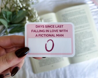 Könnte beinhalten: Eine rechteckige rosa und weiße Karte mit dem Text "DAYS SINCE LAST FALLING IN LOVE WITH A FICTIONAL MAN" und der Zahl "0" in Dunkelrosa. Die Karte wird vor einem offenen Buch gehalten.