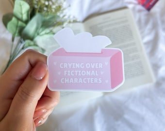 Könnte beinhalten: Ein rosa Aufkleber in Form einer Taschentuchbox mit dem Text "Crying Over Fictional Characters" in Weiß. Der Aufkleber wird von einer Hand gehalten, mit einem Buch und Blumen im Hintergrund.