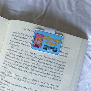May include: A blue library card bookmark with a cartoon image of a girl on the phone and the words "Library Card". The bookmark is placed on the pages of a book.