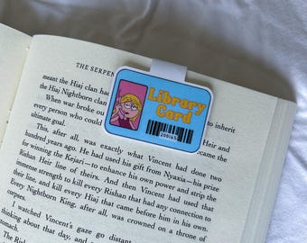 Puede incluir: Un marcapáginas azul con forma de tarjeta de biblioteca con una imagen de dibujos animados de una chica hablando por teléfono y las palabras "Library Card". El marcapáginas está en las páginas de un libro.