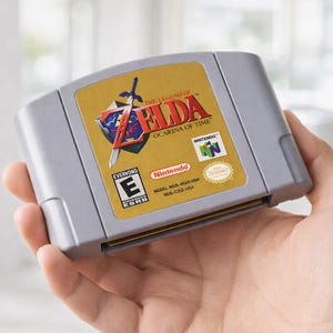 May include: A gray Nintendo 64 game cartridge for "The Legend of Zelda: Ocarina of Time." The cartridge has a gold label with the game's title, a shield, and a sword. The label also includes the Nintendo logo and an "E" rating.