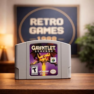 May include: A gray Nintendo 64 video game cartridge for "Gauntlet Legends" with a purple and gold label. The label features a winged demon and the game's title. The cartridge is on a wooden surface with a "Retro Games" sign in the background.