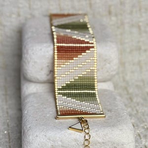 May include: A beaded bracelet with a gold-tone metal frame and chain. The bracelet features a geometric pattern in shades of white, green, and orange. The bracelet is resting on a white stone.
