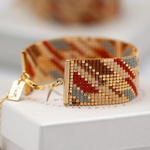 May include: A beaded bracelet with a geometric pattern in shades of gold, red, brown, and gray. The bracelet is on a white surface, with a second bracelet out of focus in the background. The bracelet has a gold-colored clasp and chain.