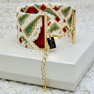 May include: Wide beaded bracelet with a festive pattern in red, green, gold, and white. The bracelet has a clasp and an extender chain. The bracelet is on a white box.