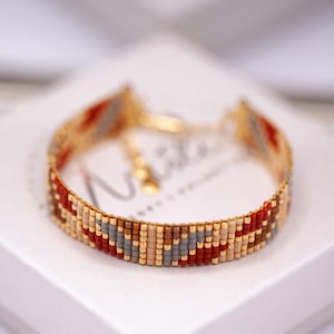 May include: A beaded bracelet with a gold-colored chain and clasp. The bracelet features a pattern of small, colorful beads in shades of red, gray, tan, and gold. The bracelet is displayed on a white surface.