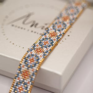 May include: A beaded bracelet with a geometric pattern in shades of blue, orange, white, and gold. The bracelet is lying on a white box with the name "Anita" printed on it. The bracelet is made of small, colorful beads.