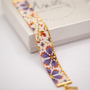 Op de afbeelding: Een kralenarmband op een witte achtergrond, met een patroon van paarse, oranje en gouden kralen. De armband heeft goudkleurige metalen uiteinden en een delicate ketting. De armband ligt op een witte doos.