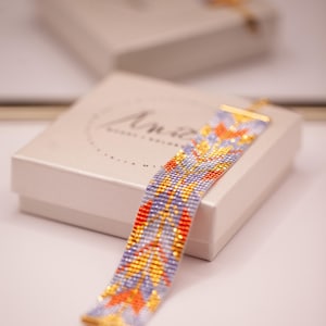 May include: A wide woven bead bracelet featuring a repeating geometric pattern in shades of light purple, bright orange, and metallic gold. The bracelet has gold-toned metal end bars and is displayed resting on a light-colored square gift box.