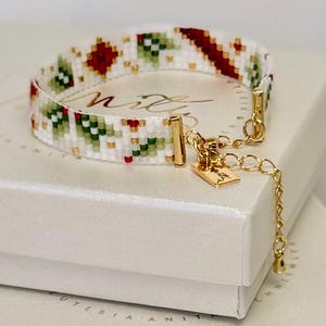 May include: A beaded bracelet with a white background and a pattern of red and green geometric shapes. The bracelet has gold-colored clasps and a small tag. The bracelet is resting on a white box.