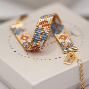 Puede incluir: Pulsera de cuentas doradas con un patrón geométrico en azul, naranja, blanco y dorado. La pulsera se muestra en una caja blanca. La pulsera tiene una cadena y un cierre. La caja tiene texto dorado.