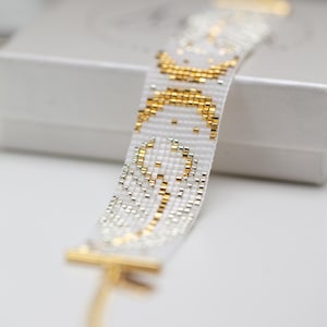 May include: White beaded bracelet with gold and silver accents. The bracelet features a repeating pattern of gold beads and silver beads, with gold clasps. The bracelet is on a white surface.