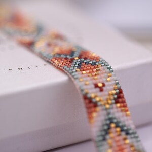 Op de afbeelding: Close-up van een kralenarmband met een geometrisch patroon. De armband heeft een mix van kleuren, waaronder oranje, teal, goud en witte kralen. De armband ligt op een wit oppervlak.