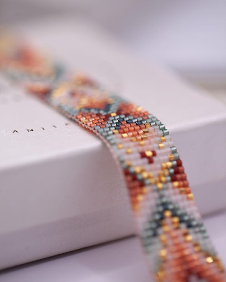 Op de afbeelding: Close-up van een kralenarmband met een geometrisch patroon. De armband heeft een mix van kleuren, waaronder oranje, teal, goud en witte kralen. De armband ligt op een wit oppervlak.