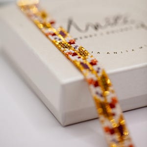 May include: A beaded bracelet with a gold clasp, featuring a pattern of white, gold, red, and purple beads. The bracelet is resting on a white box with the brand name "Anisa Anita" printed on it.