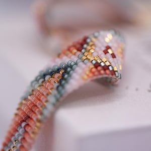 Op de afbeelding: Close-up van een kralenarmband met een kleurrijk patroon. De armband heeft een mix van kleuren, waaronder perzik, wit, goud en donkerrode kralen. De kralen zijn in een gestreept patroon gerangschikt, waardoor een textuur ontstaat. De armband is waarschijnlijk handgemaakt.
