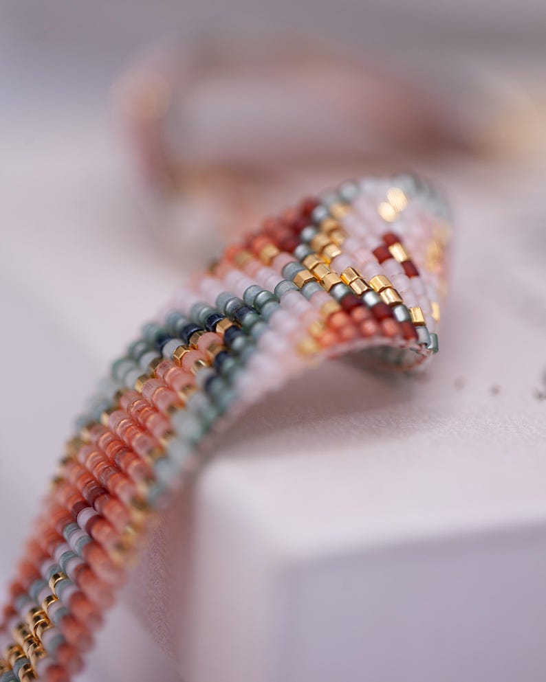 Op de afbeelding: Close-up van een kralenarmband met een kleurrijk patroon. De armband heeft een mix van kleuren, waaronder perzik, wit, goud en donkerrode kralen. De kralen zijn in een gestreept patroon gerangschikt, waardoor een textuur ontstaat. De armband is waarschijnlijk handgemaakt.