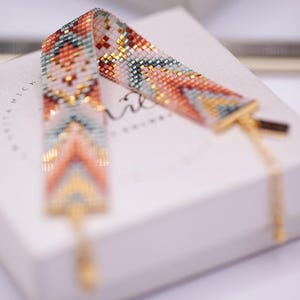 Puede incluir: Una pulsera de cuentas con un patrón geométrico en tonos melocotón, turquesa, dorado y blanco. La pulsera se muestra en una caja blanca con herrajes dorados y una delicada cadena.