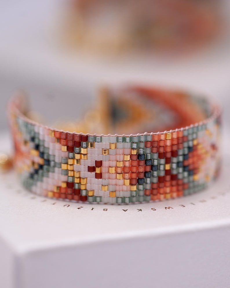 Op de afbeelding: Een kleurrijke kralenarmband met een geweven patroon. De armband heeft een mix van oranje, rode, gouden, groene en witte kralen. De armband ligt op een wit oppervlak.