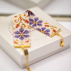 May include: A beaded bracelet with a floral pattern in white, purple, orange, and gold. The bracelet is resting on a white box. The bracelet has a gold-coloured clasp and chain. The text "ANITA WIEKO" is visible on the box.