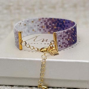 May include: A beaded bracelet with a gradient of white, lavender, and purple hues. The bracelet features gold-coloured metal accents and an adjustable chain. The beads have small gold-coloured accents. The bracelet is displayed on a white box.