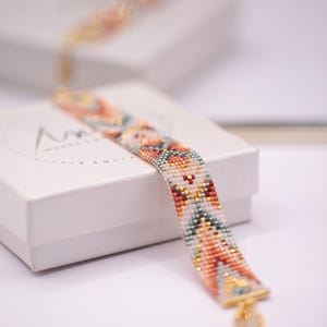 Op de afbeelding: Een kralenarmband met een kleurrijk geometrisch patroon in tinten oranje, teal, goud en wit. De armband ligt op een witte doos. De armband heeft een gouden sluiting.