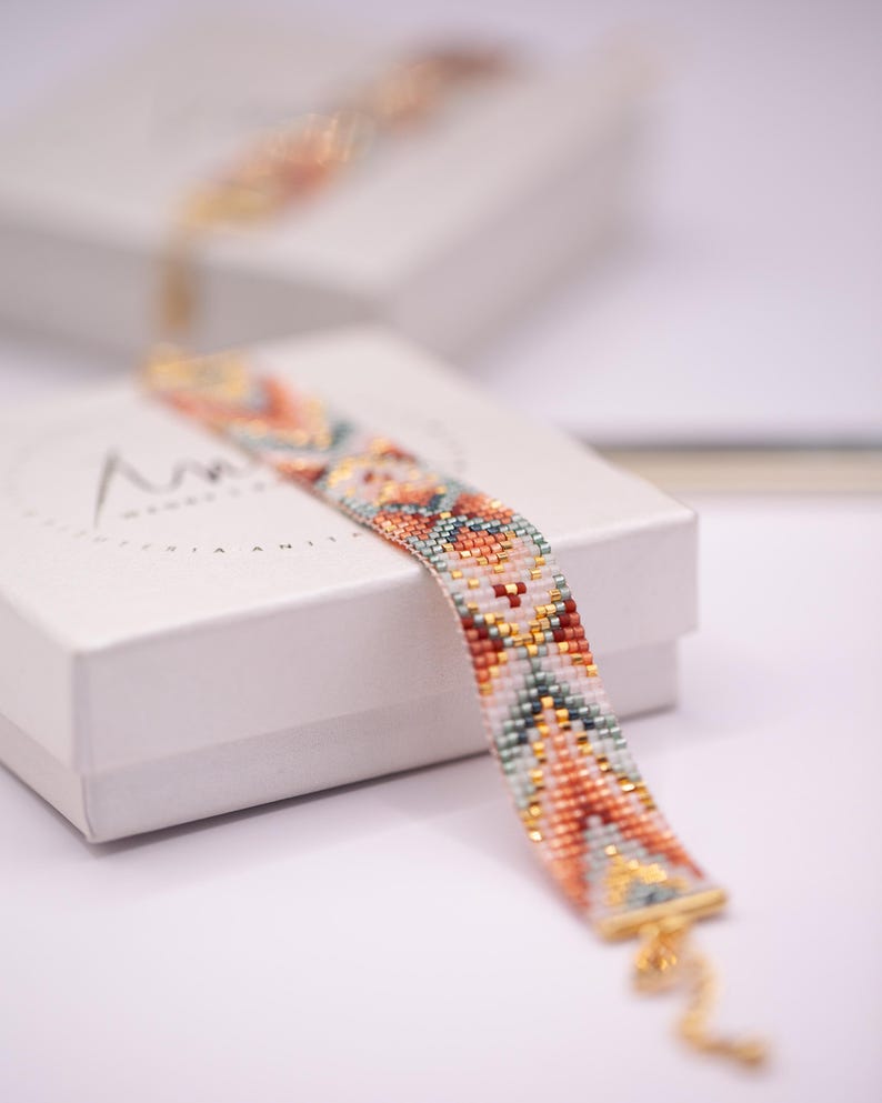 Op de afbeelding: Een kralenarmband met een kleurrijk geometrisch patroon in tinten oranje, teal, goud en wit. De armband ligt op een witte doos. De armband heeft een gouden sluiting.
