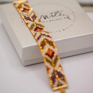 May include: A beaded bracelet with a geometric pattern in gold, white, purple, orange, and red. The bracelet is displayed on a white surface with a box in the background. The box has the text "Nita Michn" in a circular design.